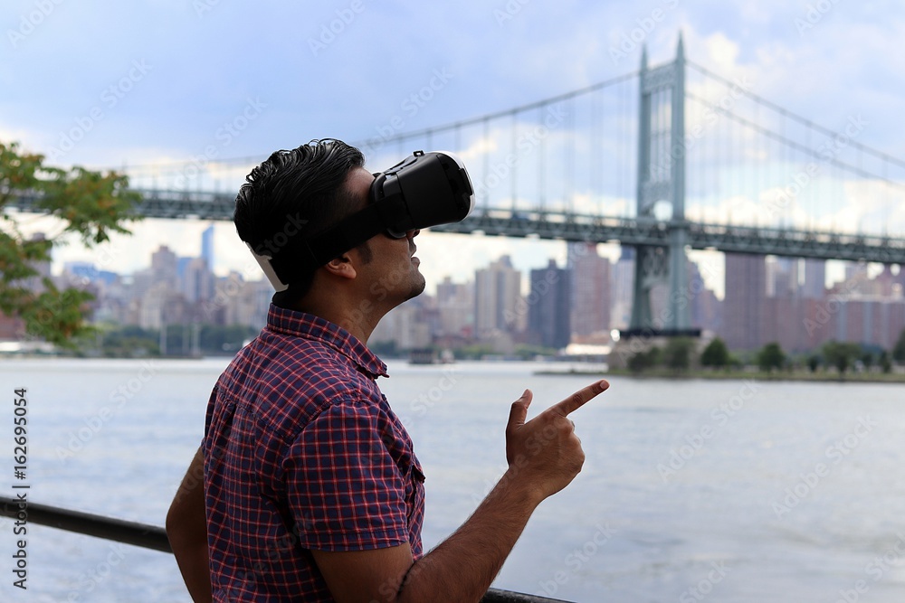Young handsome man wearing virtual reality goggles headset while standing infont of waterfront bridge and urban city skyline
