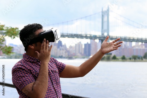 Young handsome man wearing virtual reality goggles headset while standing infont of waterfront bridge and urban city skyline photo