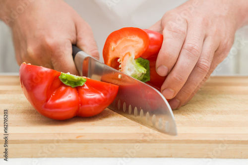 Chef's hands with knife cutting sweet paprika on the wooden board. Preparation for cooking. Healthy eating and lifestyle.