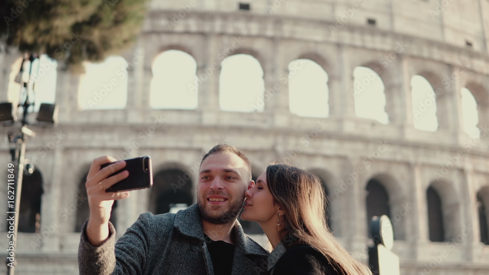 Young attractive woman and man standing near the Colosseum in Rome, Italy. Couple takes the selfie photo on smartphone.