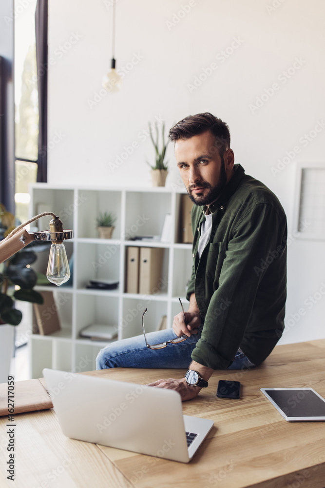 Handsome bearded man sitting on table with digital devices and looking at camera Handsome bearded man sitting on table with digital devices and looking at camera