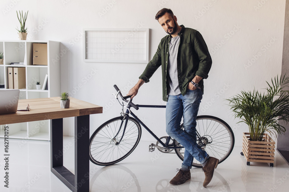Handsome bearded man standing with bicycle and looking at camera in office Handsome bearded man standing with bicycle and looking at camera in office