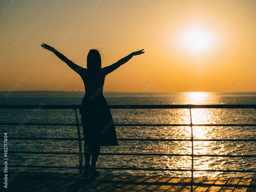 Silhouette of the happy woman in summer dress standing at the beach during sunset. Natural light and colors