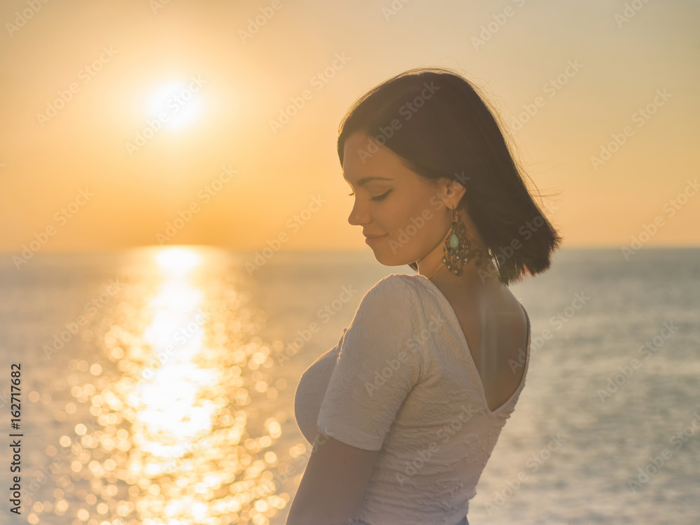 Portrait of young attractive woman standing in morning sunlight close to sea or ocean. Beautiful lady with short brunette hair and long earrings.