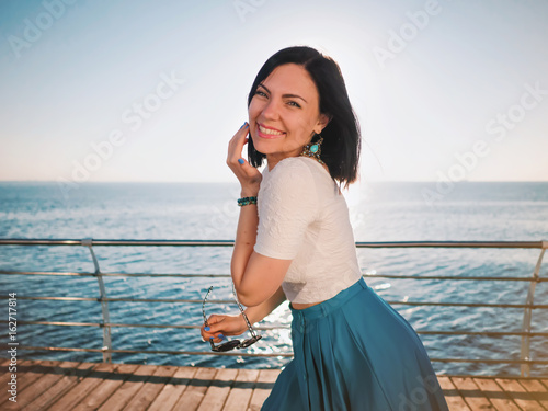 Beautiful young woman posing as model standing on embankment near sea or ocean. Lady wearing retro style skirt and sunglasses. Morning sunlight.
