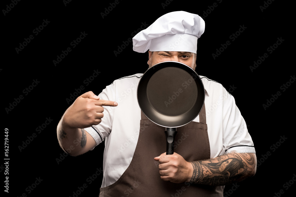 professional chef in hat and apron winking and pointing on frying pan isolated on black professional chef in hat and apron winking and pointing on frying pan isolated on black