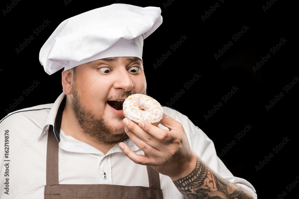 Excited chef in hat and apron eating doughnut isolated on black Excited chef in hat and apron eating doughnut isolated on black