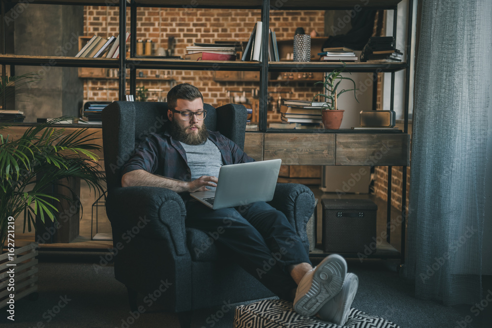 Young bearded man in eyeglasses using laptop while sitting in armchair at home