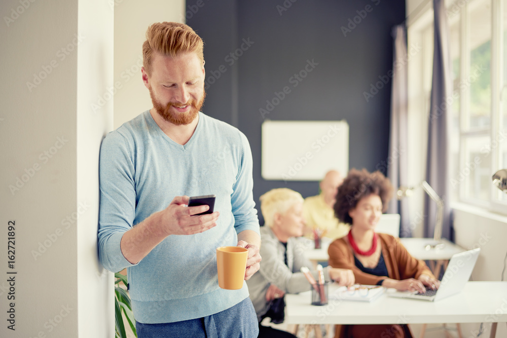 Young Caucasian businessman using smartphone while standing and drinking coffee in modern office Young Caucasian businessman using smartphone while standing and drinking coffee in modern office