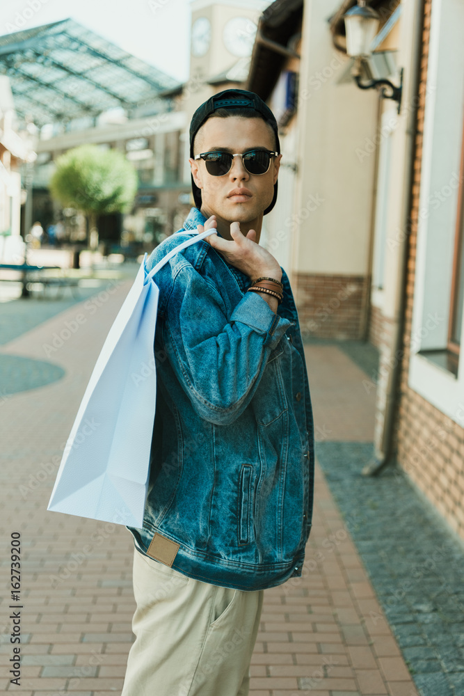 Fototapeta premium young stylish man in sunglasses holding shopping bag and standing on the street