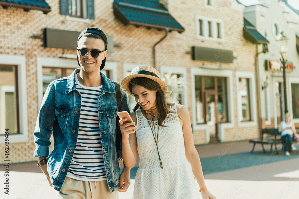 Smiling young woman in straw hat using smartphone while standing near handsome boyfriend outdoor