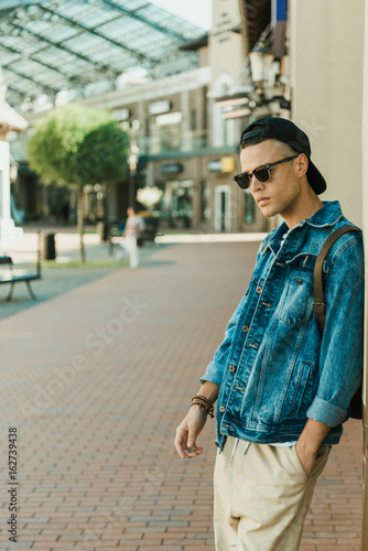 Stylish young man in denim jacket and sunglasses standing outdoor looking away
