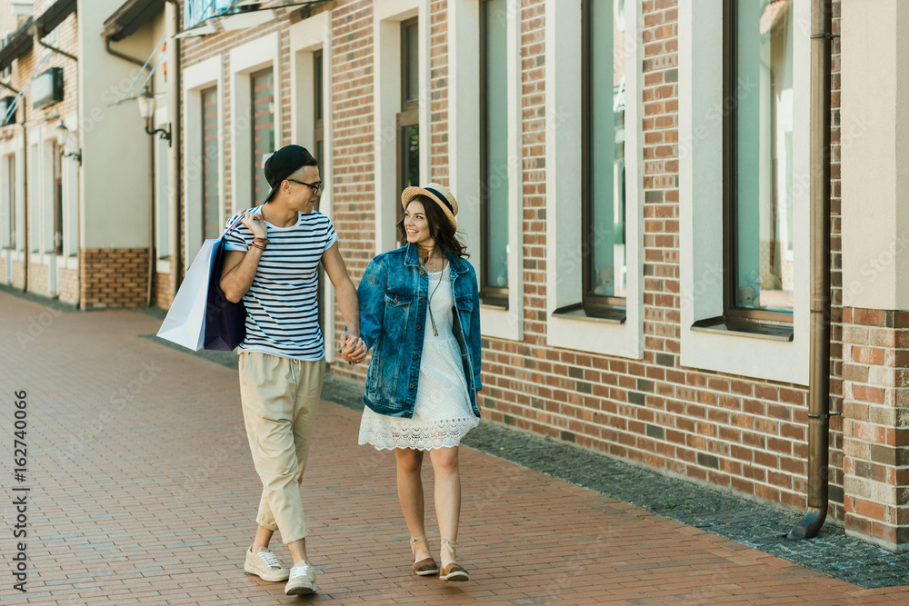 happy young stylish couple with shopping bags holding hands and walking in city