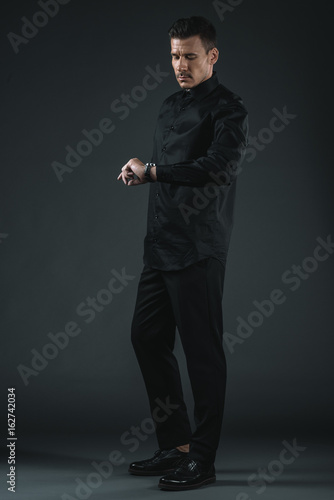serious fashionable man in black outfit looking at watch, isolated on grey