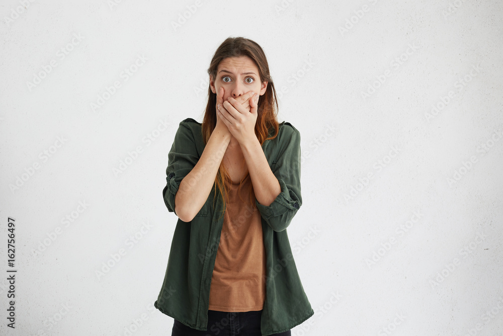 Shocked woman with long dyed hair wearing green clothes looking with bugged eyes covering her mouth with hands looking into camera with despair trying to keep silence and not telling secret to anybody Shocked woman with long dyed hair wearing green clothes looking with bugged eyes covering her mouth with hands looking into camera with despair trying to keep silence and not telling secret to anybody