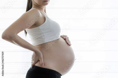 Pregnant woman holding back in front of white curtain