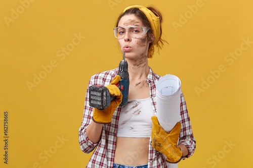 Self-confident female builder wearing goggles, white top and checkered shirt, protective gloves holding drill and papers being dirty after hard work isolated over yellow background. Maintenance photo