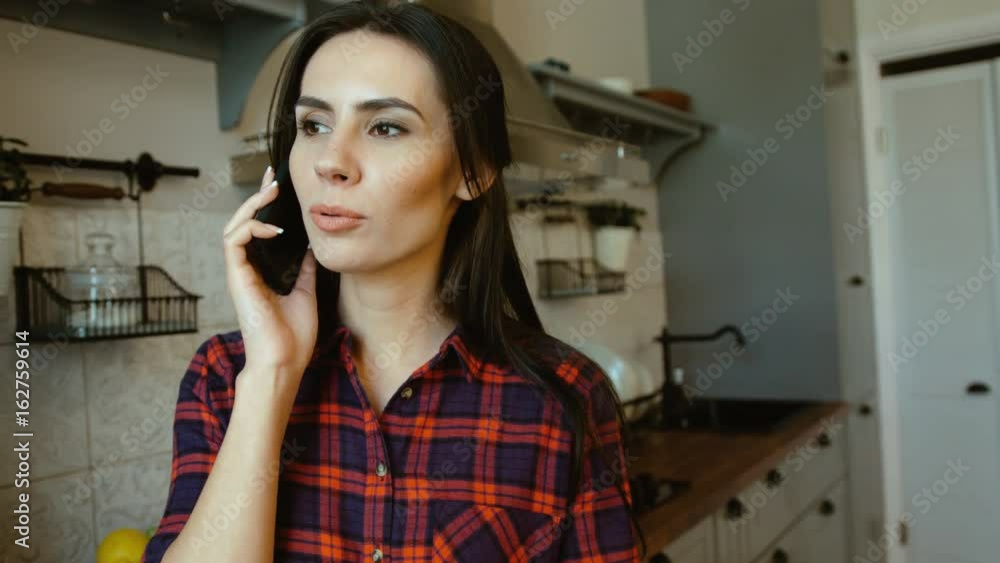 Attractive woman talking on the smart phone whikle standing in her kitchen. Woman having conversation. Close up Attractive woman talking on the smart phone whikle standing in her kitchen. Woman having conversation. Close up