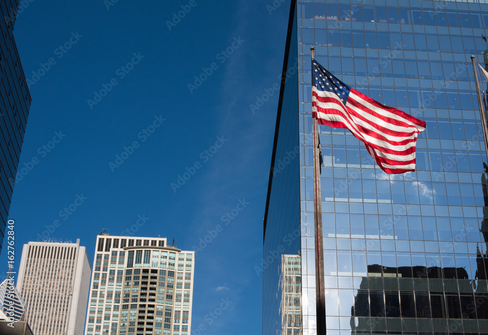 American flag rising above a city plaza, with skyscrapers behind, reflecting a beautiful blue sky