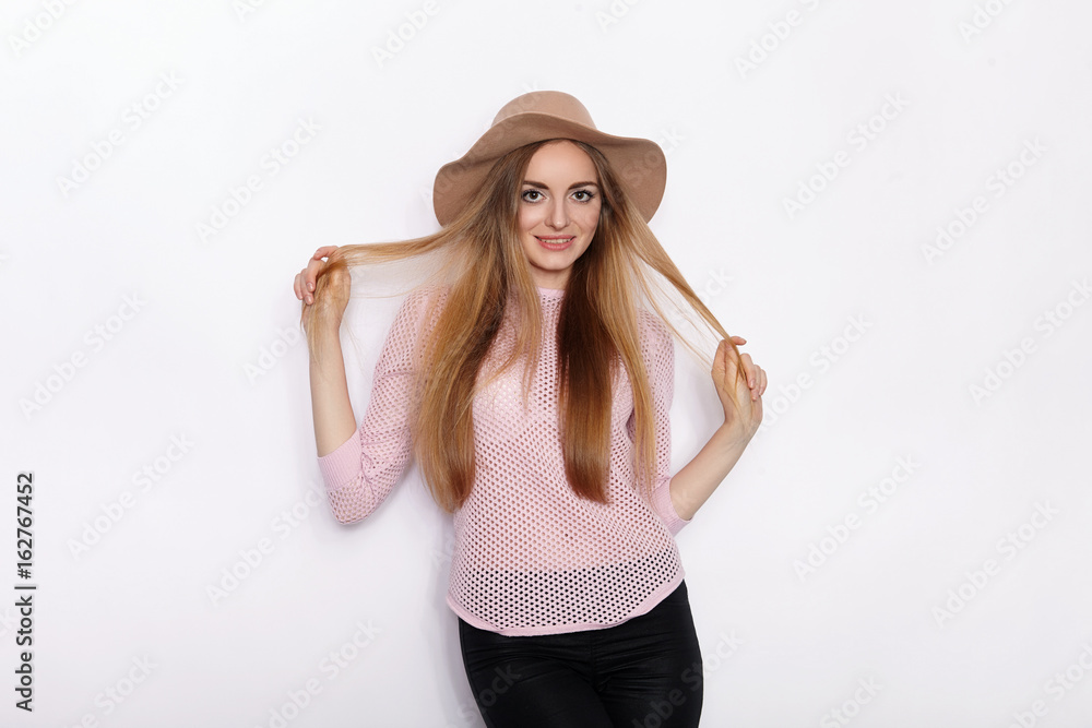 Cute and stylish beautiful young blonde woman in trendy clothing practicing model poses and looking away with smile while standing against white wall background Cute and stylish beautiful young blonde woman in trendy clothing practicing model poses and looking away with smile while standing against white wall background