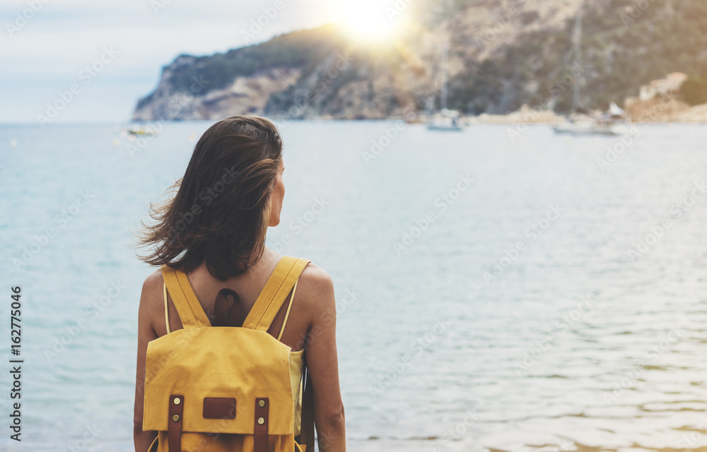 Fototapeta premium Back view hipster girl with backpack in sand coastline on nature landscape, mock up. Traveler on background beach seascape and horizon mountain. Tourist look on blue sun ocean, summer relax lifestyle