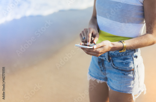 Hipster girl hold on smart phone gadget in sand coastline, mock up of blank screen. Traveler using in female hand mobile on background beach seascape horizon. Tourist look on blue sun ocean, lifestyle