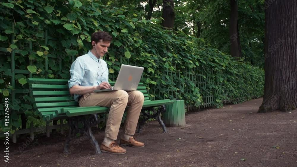 Caucasian young man using laptop with headphones sitting on bench at city park. Summer day. Freelancer working outdoors. Trees and green spaces at background. Caucasian young man using laptop with headphones sitting on bench at city park. Summer day. Freelancer working outdoors. Trees and green spaces at background.