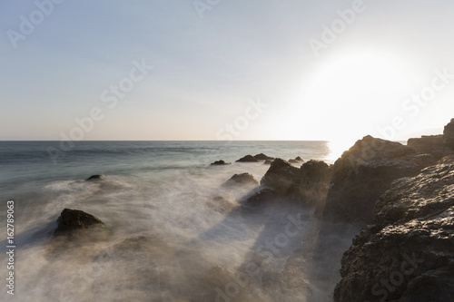 Motion blur waves breaking over rocks with setting sun at Pirates Cove in Malibu, California.