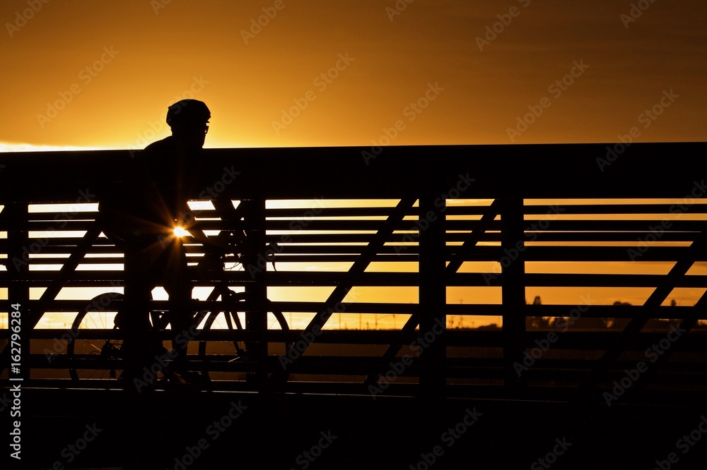 Φωτογραφία Cyclist Crossing a Bridge  - a silhouette of a male cyclist crossing a foot/bicycle bridge during evening light with a single starburst of sunlight shining through, near Fresno, California, U