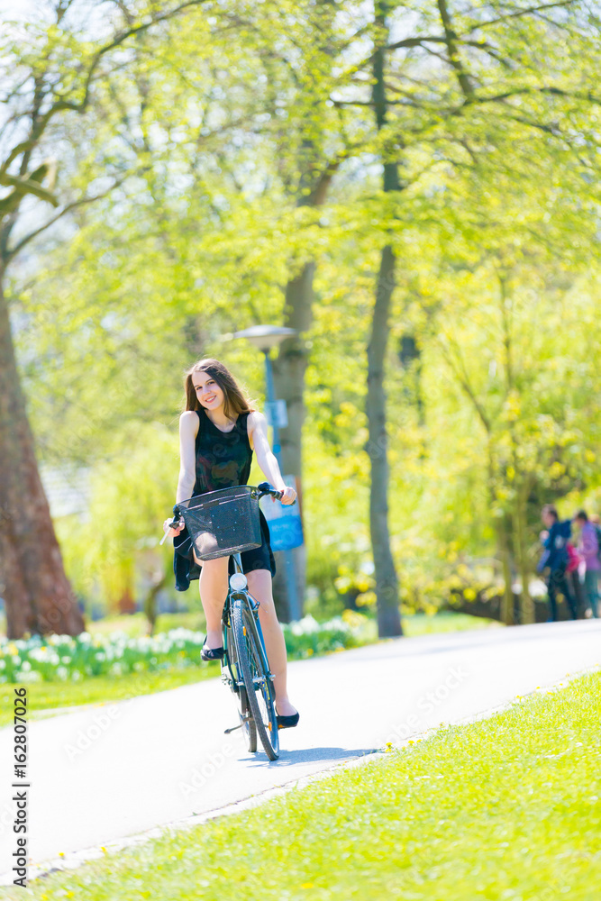 Portrait view of girl on bicycle wearing on black short dress. Young happy Woman riding along road on green spring outdoor Park. Sporty young girl riding a bicycle on a sunny morning, view from face Portrait view of girl on bicycle wearing on black short dress. Young happy Woman riding along road on green spring outdoor Park. Sporty young girl riding a bicycle on a sunny morning, view from face