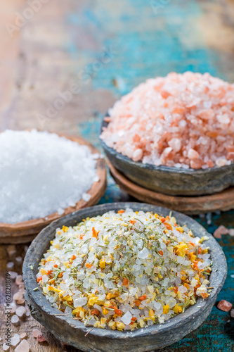 Three different types of natural salt in stone bowls on wooden surface. White sea salt, pink Himalayan salt, Spiced salt with rosemary photo