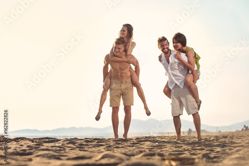 Two young men giving their girlfriends piggyback rides at the beach. Cheerful young friends enjoying summertime on the beach.
