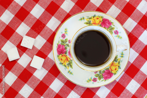 Black tea in vintage porcelain cup, sugar cubes on red and white checkered cloth
