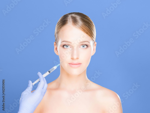 Doctor making injections in a beautiful face of a young woman over blue background. Plastic surgery concept.