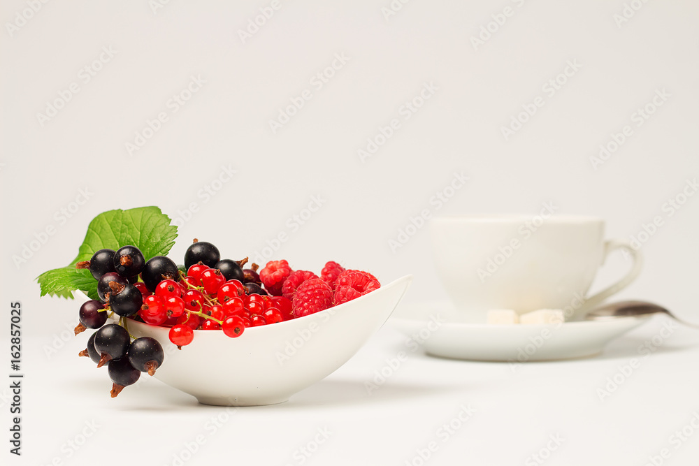 Ripe fresh black and red berries in a porcelain saucer. Currant and raspberry on a branch with leaf. Cup of coffee with milk in the background Ripe fresh black and red berries in a porcelain saucer. Currant and raspberry on a branch with leaf. Cup of coffee with milk in the background