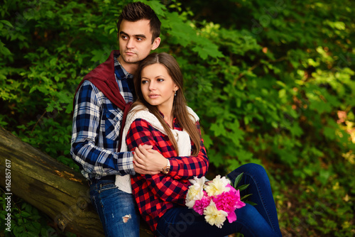 Stylish young couple in shirts and jeans while walking in the forest. A beautiful girl with her handsome boyfriend in a hug sitting on a fallen tree.