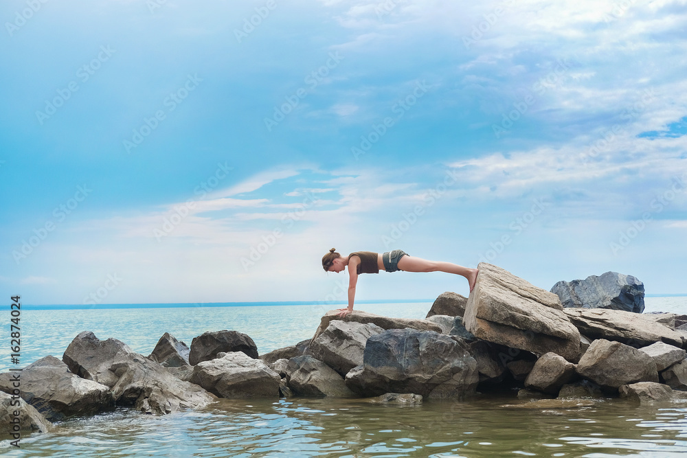 Girl practicing yoga on the rocks against the blue sky and the azure sea. Woman stands on a stone in a plank posture.