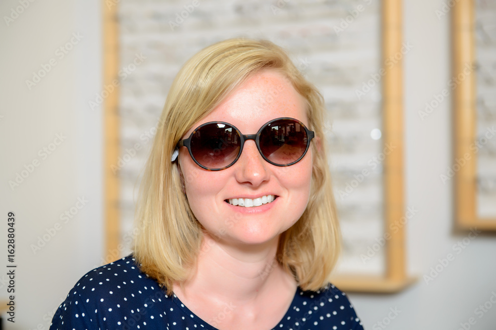 Naklejka premium in optician shop- young woman selecting new sunglasses