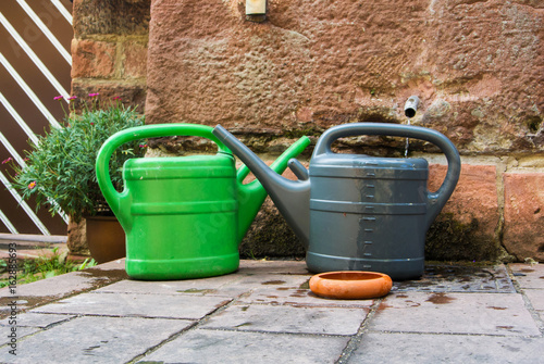 Two plastic watering cans, green and gray, ready for pour the garden standing on the stone pavement near the wall, Heidelberg, Germany. © Victoria