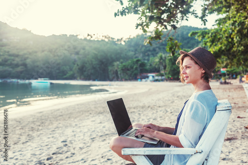 Young beautiful woman hipster traveler freelancer with laptop on the seashore. Business and vacation concept