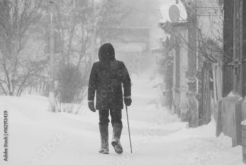 Lonely elderly man with a cane walking leisurely through a snow storm through the streets of the city