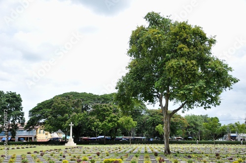 big trees and lawn in a cemetery with headstones in the background at Kanchanaburi War Cemetery in Thailand.