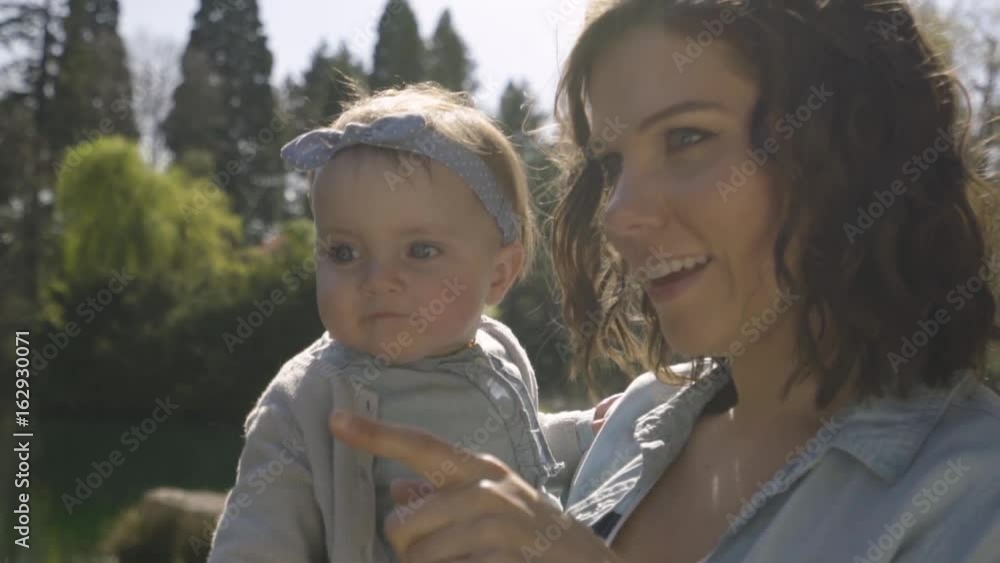 Happy Mother Holds Cute Baby Girl And Points To Something In Distance In Park On Sunny Day Happy Mother Holds Cute Baby Girl And Points To Something In Distance In Park On Sunny Day