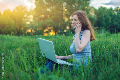 Woman sitting on a green meadow on the background of sunset with clouds. Working on laptop and talking on the phone.