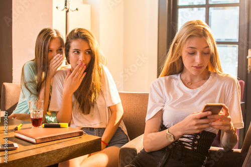 Teenage girl texting on mobile phone with her friends gossiping at the background.