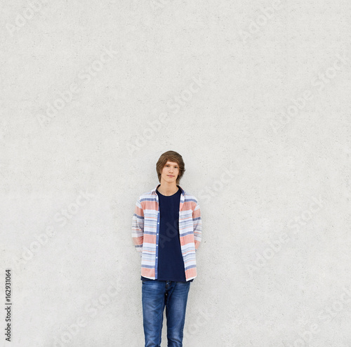 Young boy with trendy haircut wearing shirt and jeans standing near white concrete wall having serious expression. Stylish teenger posing into camera while being isolated over white background photo