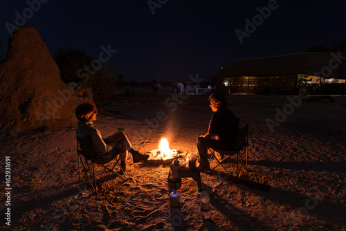 Couple sitting at burning camp fire in the night. Camping in the desert with wild elephants in background. Summer adventures and exploration in the african National Parks.
