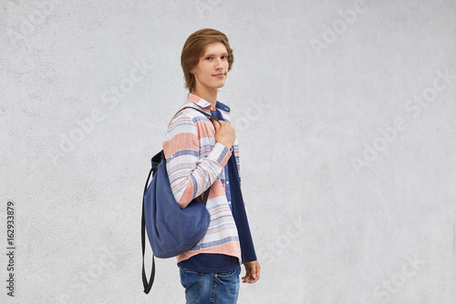 Teenage boy standing sideways holding rucksack wearing shirt and jeans posing against white concrete wall with copy space for your advertisment or promotional text. Boy with backpack going to school
