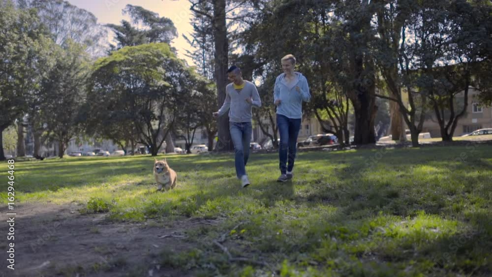 Gay Couple Run Through Field In Park, With Their Adorable Corgi Dog Gay Couple Run Through Field In Park, With Their Adorable Corgi Dog