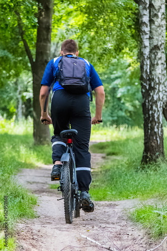 A seventeen-year-old boy, a teenager, rides a bicycle through the forest in the mountains. He is wearing a blue T-shirt and black sports pants, with a backpack behind his shoulders.